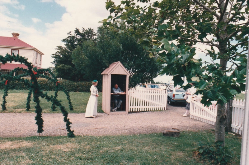 Christmas, past and present at Howick Historical Village, 12 December 1987. A woman in costume is standing next to a man in a sentry box by the gate. Greenery hoops are over the road.