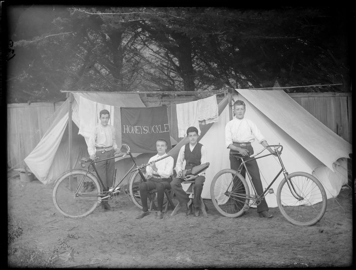 Four unidentified boys, two with bicycles and two with flute and autoharp, in front of two tents with washing line between with 'Honeysuckle' banner, wooden fence and trees beyond, [Sumner?], Christchurch