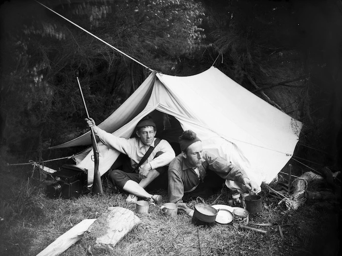 L H Duval and William Williams sitting outside a tent with a gun, camera and other equipment