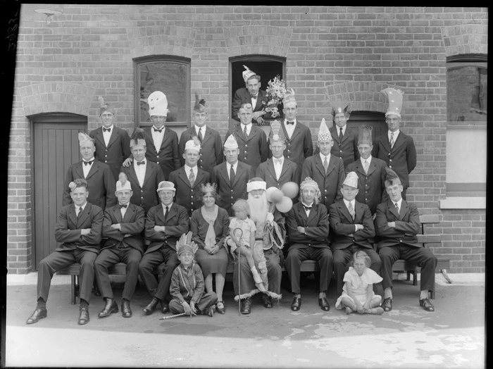 Unidentified group, wearing party hats, outside a brick building, probably Christchurch district, includes man dressed up as Santa Claus holding balloons, with a small child sitting on his lap