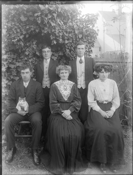 Family portrait in the backyard with wooden fence behind, unidentified older woman with large lace collar with three men, a woman and a cat, probably Christchurch region