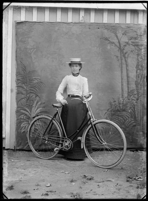 Outdoors portrait in front of false backdrop, an unidentified young woman with straw hat, black neck ribbon and a women's style bicycle, probably Christchurch region