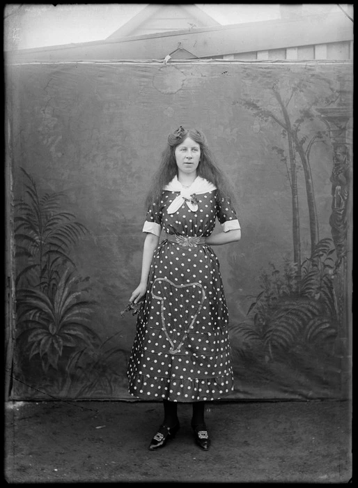 Outdoors portrait in front of false backdrop, an unidentified young woman wearing polka dot harp pattern dress, scarf and buckled shoes, with flowers in hand, probably Christchurch region