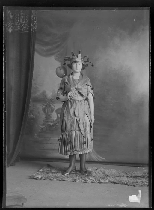 Studio portrait of unidentified young woman dressed as a jester, with balloon on a stick, dress and crown with pompoms, Christchurch