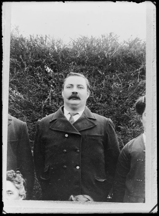 Outdoors upper torso portrait of unidentified older man with large moustache and double breasted overcoat in front of tall hedge, people surrounding, probably Christchurch region