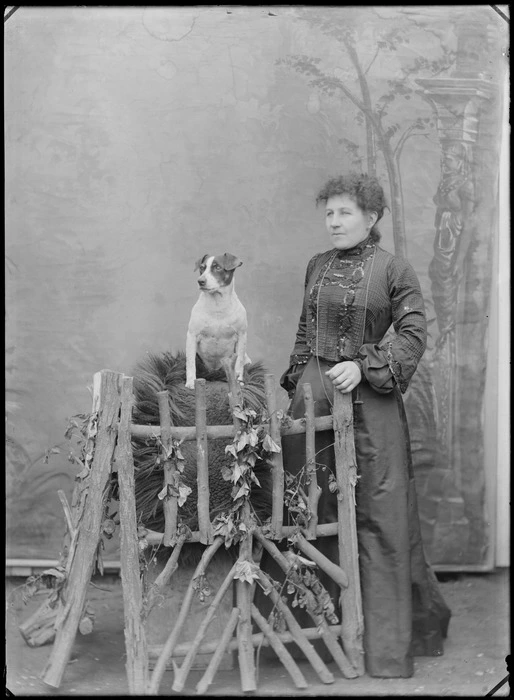 Outdoors portrait in front of false backdrop, unidentified woman with high neck collar dark embroidered blouse, standing with old Jack Russell Terrier dog, probably Christchurch region