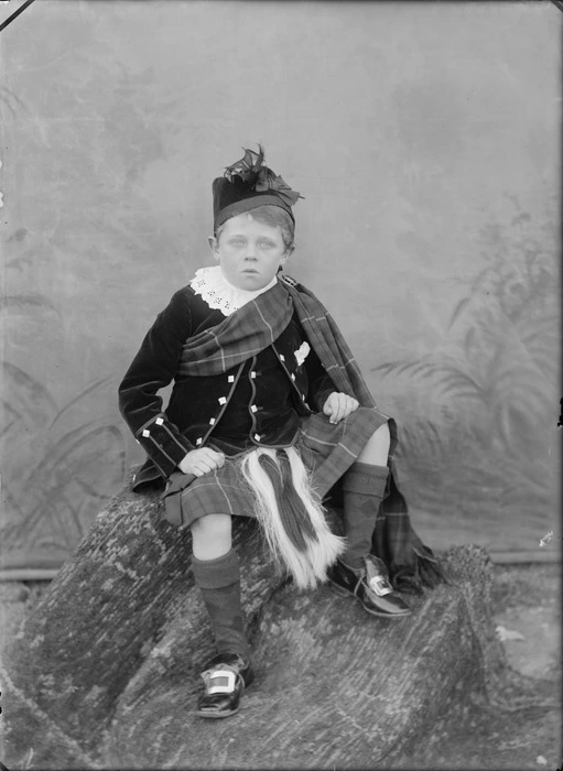 Outdoors portrait in front of false backdrop, an unidentified young boy in Scottish clansman costume, jacket and kilt with piper's sporran, lace collar, tartan sash with brooch and Glengarry hat, Christchurch
