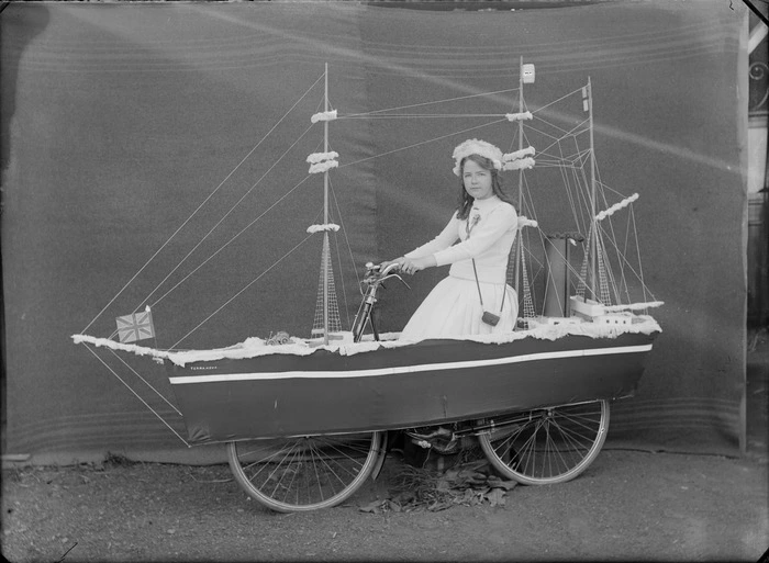 Outdoors portrait in front of false backdrop, an unidentified young woman with rosette, on a bicycle float of Robert Falcon Scott's boat the 'Terra Nova', probably Christchurch region