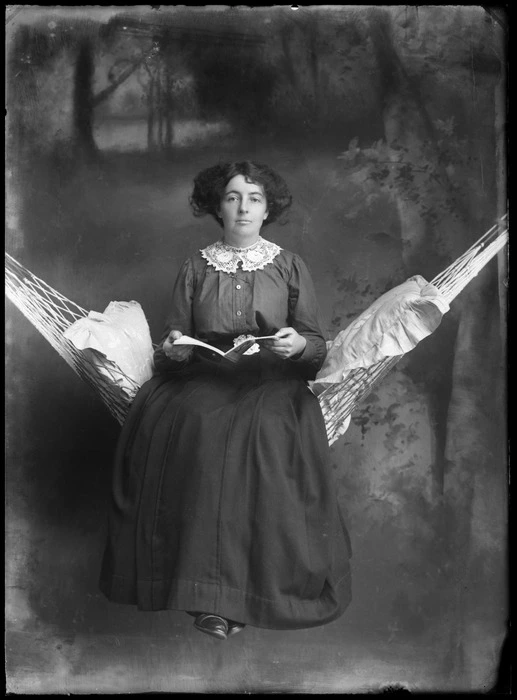 Studio portrait of unidentified woman, sitting on a hammock with cushions and reading a book, probably Christchurch district