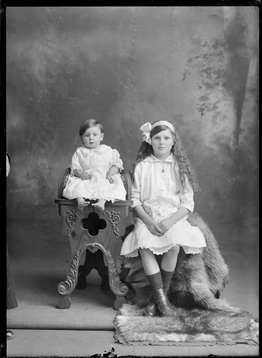 Studio family unidentified portrait, older sister with long hair, bow and locket sitting on fur rug next to toddler sister in lace collar cotton dress on wooden high chair, Christchurch