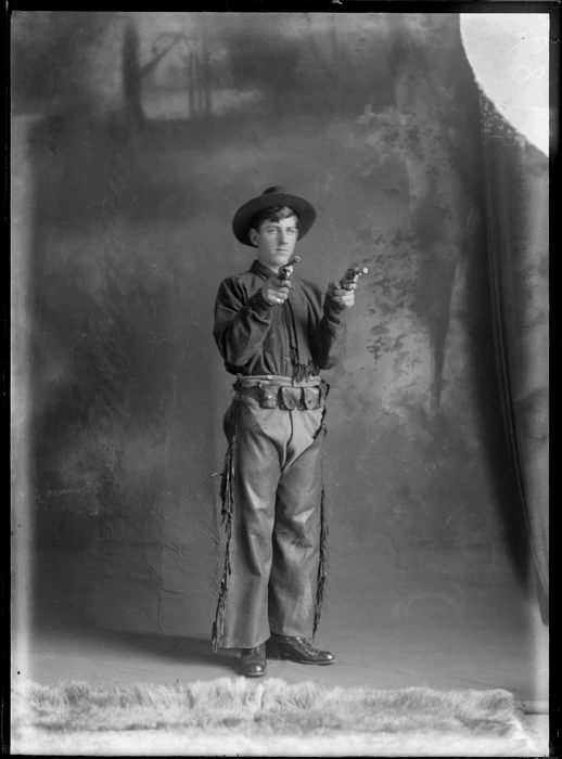 Studio portrait of unidentified young man dressed as a cowboy, with leather chaps with tassels, dark shirt and scarf, army ammunition belt, hat, and holding two toy guns, Christchurch