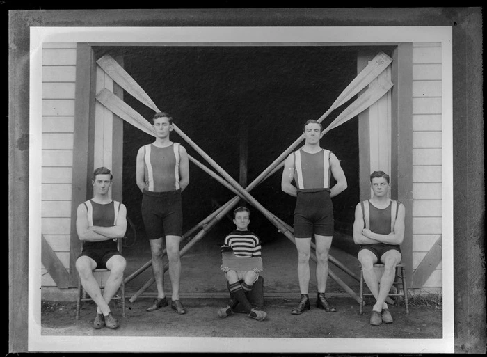 Group portrait of members of the Avon Rowing Club, showing four young men in swimming costumes, with a young boy coxswain sitting in center [holding a boat rudder?], and oars crossed diagonally behind, outside the Avon Rowing Club boatshed, Christchurch