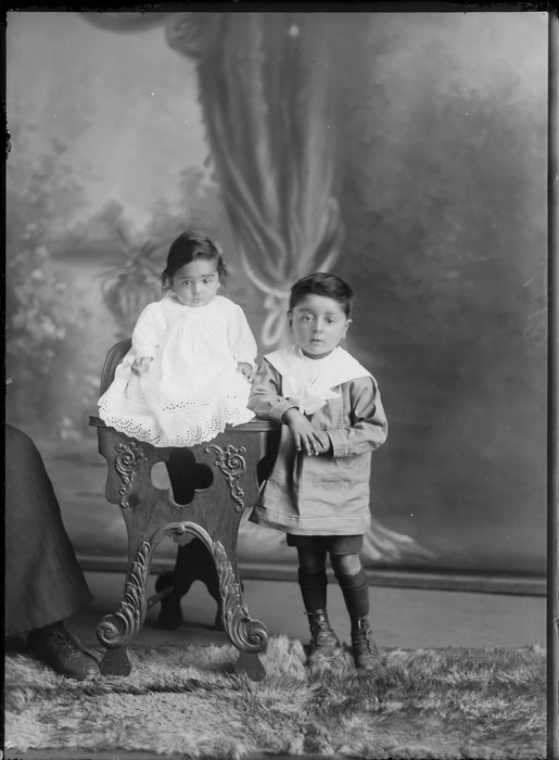 Studio unidentified family portrait, young boy with large bow collar standing next to baby sister in lace dress on wooden high chair, Christchurch