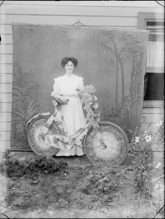 Unidentified woman outside a wooden house, with a decorated bicycle with flowers, and a painted studio backdrop, probably Christchurch district