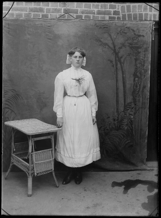 Unidentified woman, outside a brick building, wearing a ribbon, with a painted studio backdrop, probably Christchurch district