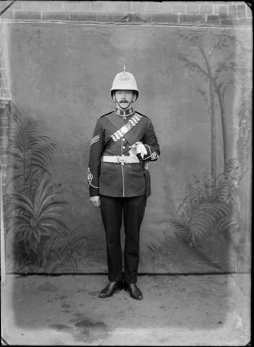 Outdoors in front of a false backdrop, portrait of an unidentified World War I sergeant in full dress uniform [Rifleman?] and swagger stick, probably Christchurch region