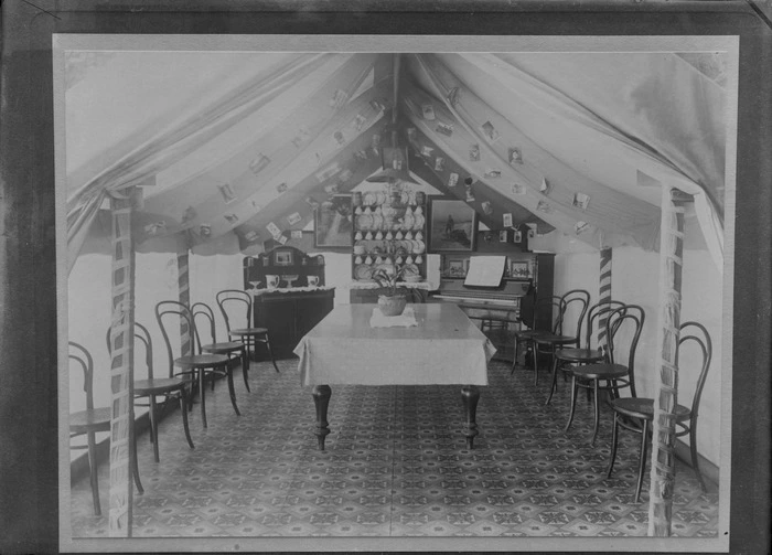 Inside a large tent setup as a dining room, with carpet, a large table with potted plant, chairs, piano, china cabinet with china and sideboard, postcards and framed pictures, [Sumner?], Christchurch
