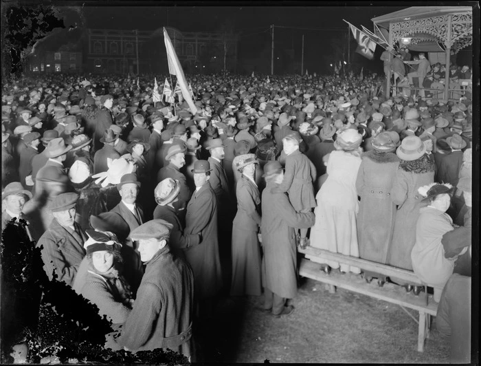 Costumes, view of people at a night rally [Marine Parade, Napier?], looking to the band rotunda with army brass band and guests, people waving flags, Hawke's Bay District