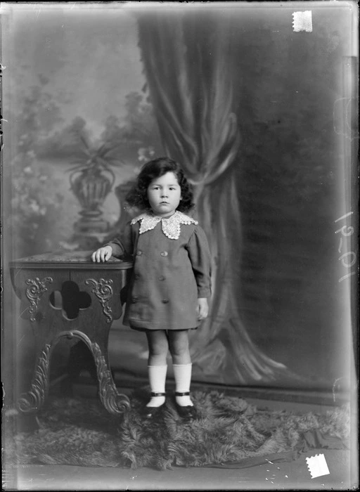 Studio portrait of unidentified young girl in a double breasted coat with a large lace collar standing next to a wooden high chair, Christchurch