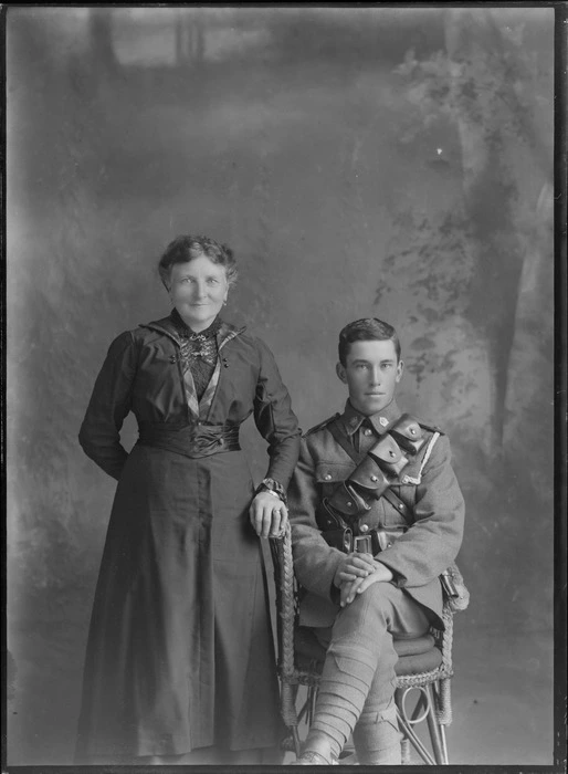 Studio unidentified family portrait of older mother standing in a dark lace blouse and dress next to her young adult son in World War I soldier's uniform with bandolier and waist ammunition belts, collar badges and white shoulder pocket cord sitting, Christchurch