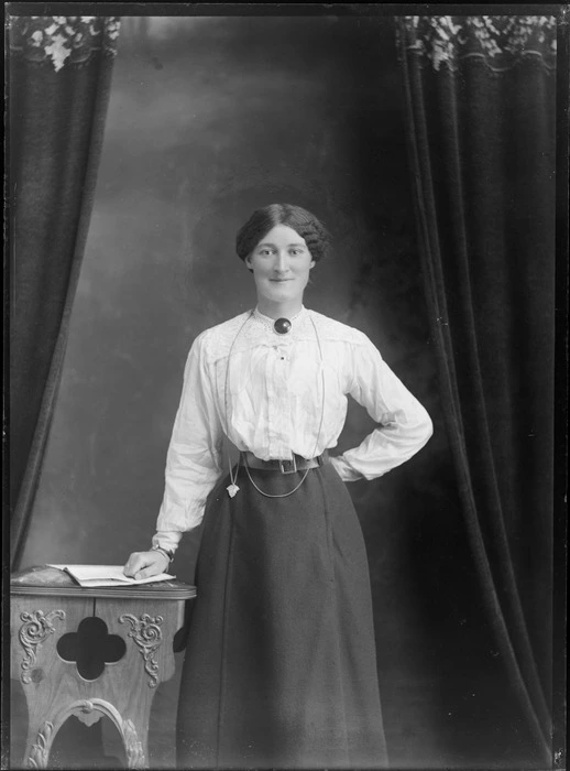 Studio unidentified portrait of a woman in a lace shoulder cotton shirt with a large circular neck brooch, dark skirt and neck chain pendant, standing with a wooden highchair and book, Christchurch