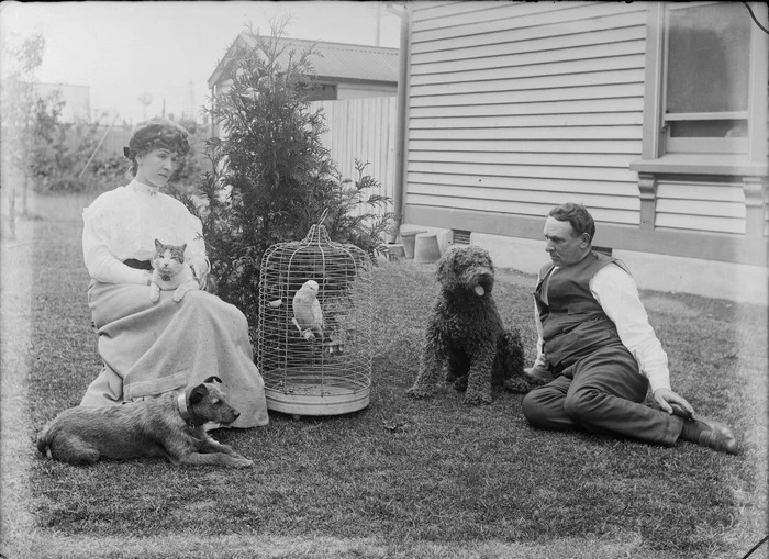 Unidentified man and woman with domestic pets, including two dogs, a cat, and a bird in a cage, sitting on a grass lawn outside a wooden building, possibly Christchurch district