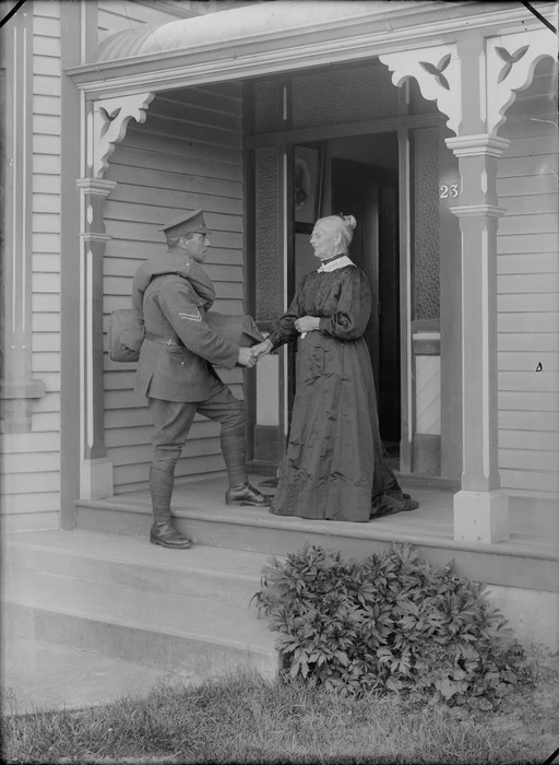 Outdoors on a veranda of a wooden house, a farewell between an unidentified elderly mother with her older World War I Corporal Soldier son carrying his coat and kit bag, probably Christchurch region