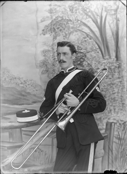 Outdoors in front of false backdrop, an unidentified portrait of a young World War I soldier with moustache in dress uniform with badges and insignia, white shoulder sash, with braided pants and flower dome hat, holding a trombone [Army Band?], probably Christchurch region
