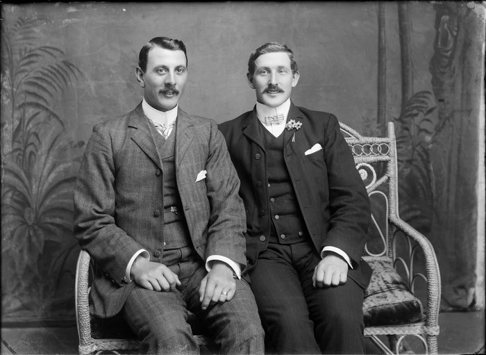 Studio portrait of two unidentified men with moustaches, sitting on a cane couch, one with lapel flowers, Christchurch