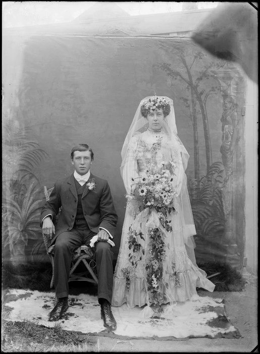 Outdoors portrait of unidentified wedding couple in front of a false backdrop, groom sitting in a three piece pinstriped suit with white bow tie and lapel flowers, bride standing with long veil and pearl necklace holding flowers, probably Christchurch region