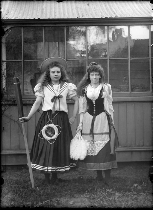 Outdoors portrait of two unidentified young women in fancy dress costumes, one as a sailor with a small 'City of Crafton, Sydney' lifebuoy and oar, and the other as a [gypsy or milkmaid?], probably Christchurch region