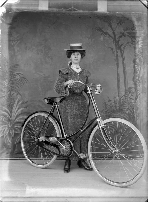 Outdoors portrait of unidentified young woman in a woollen jacket with shoulder pads and matching skirt, pearl necklace and straw hat, standing with a woman's bicycle in front of false backdrop, probably Christchurch region