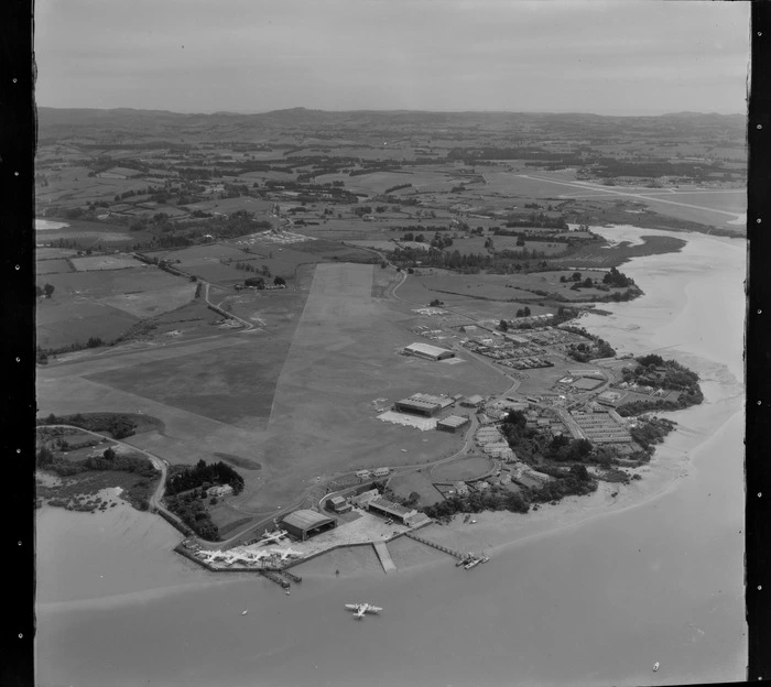 Hobsonville Aerodrome, Auckland, showing a seaplane on the ocean, with five aircrafts on shore on the tarmac next to the hangar
