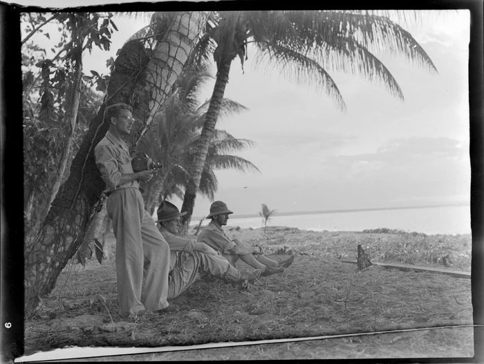 Three unidentified members of the RNZAF (Royal New Zealand Air Force), one holding a camera, under palm trees on the Guadalcanal coast, at Camp Kiwi, Solomon Islands