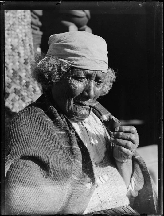 Portrait of an unidentified elderly Maori woman with a moko, bandana and pipe, sitting in front of a meeting house, probably Taupo