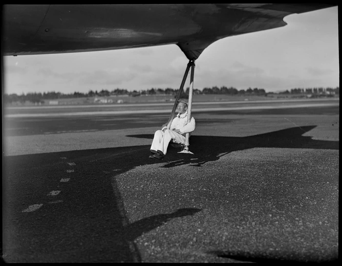 Unidentified airport staff member, swinging on tail stanchion, attached to back of aircraft VH-TAD McDouall Stuart, TAA (Trans Australia Airlines), Whenuapai Air Base, Auckland