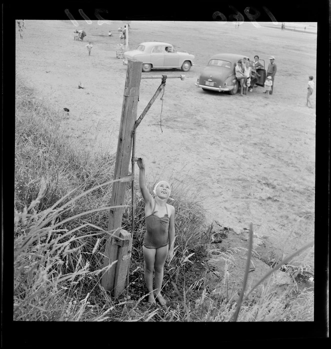 Girl turning tap on public shower, with cars and families on beach, probably Titahi Beach, Wellington