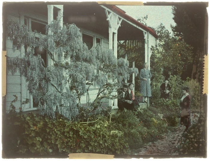 Group of unidentified women and man, standing on a wooden verandah with a wisteria vine in foreground, location unidentified