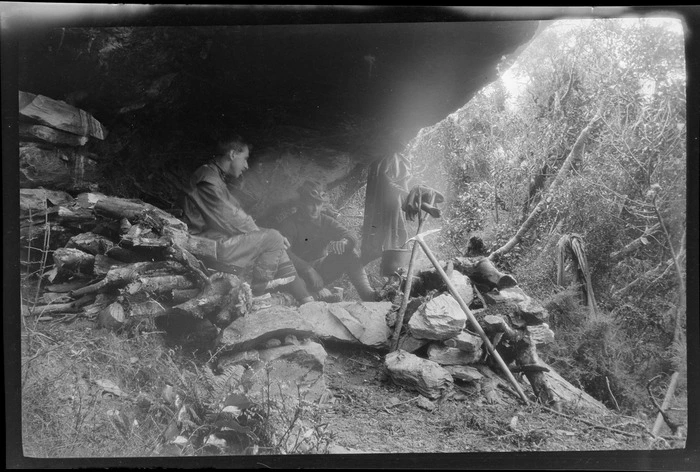 Edgar Williams and an unidentified man, sitting by a campfire under a rock overhang, [Franz Josef, Westland District?]