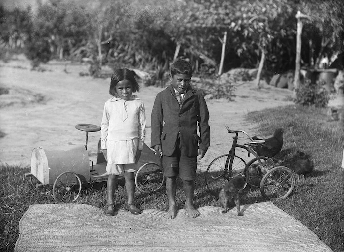 Maori girl and boy alongside a whariki, peddle-cart and tricycle
