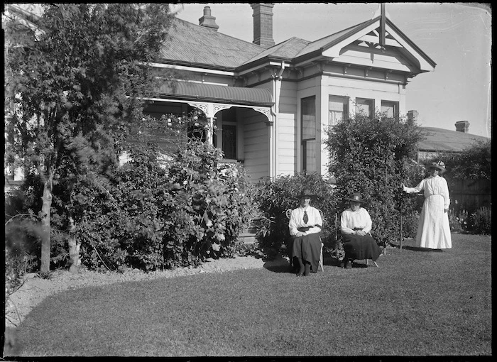 Three unidentified women in a garden, beside a villa with verandah partially visible.