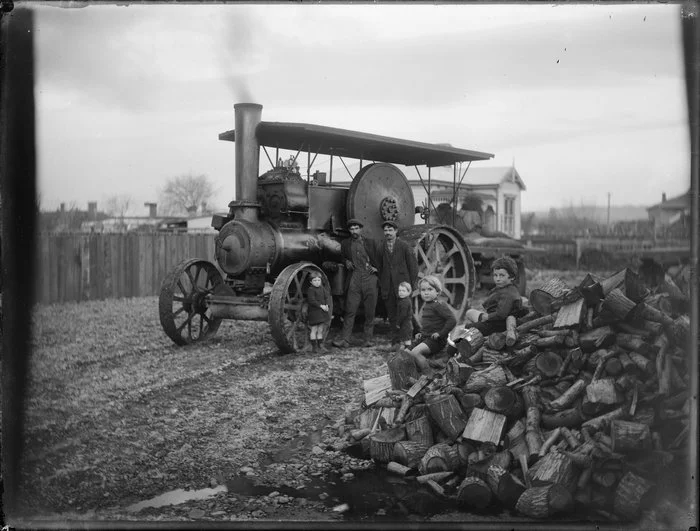 Children and men beside a large traction engine with two boys sitting on a large stack of firewood