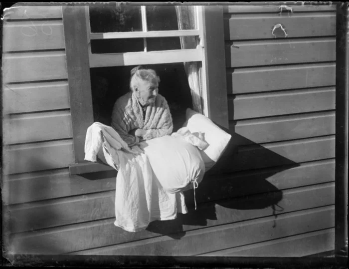 An unidentified old woman in a woolen cardigan leans out of a house window with items of bedding, including a pillow and a nightdress, location unknown