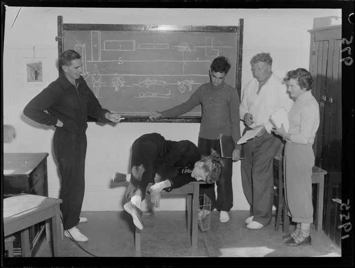 Stretch demonstrated by young woman on desk at National Amateur Athletic Coaching School, including diagrams on blackboard and with teacher and students looking on