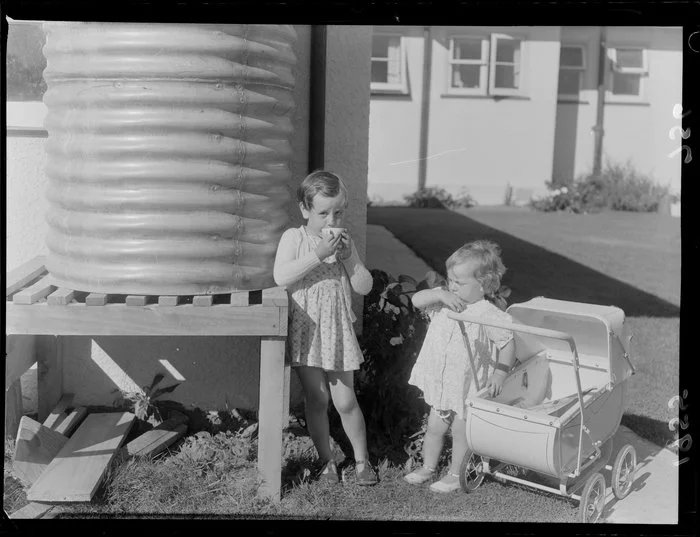 Little girl stands with another child by a backyard water tank and drinks from a cup, during a water shortage, Marton, Rangitikei District