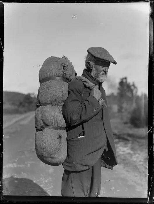 Unidentified elderly man [tramp?] with swag on his back, rural road, location unknown