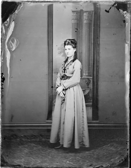 Unidentified woman with hair coiled on top of her head, wearing a dress with long sleeves and full skirt and an embroidered bodice