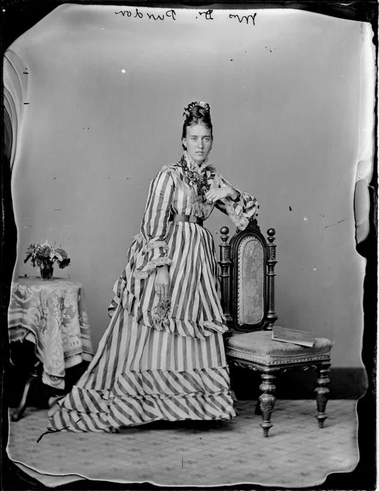 Mrs Dr Purdon, with her hair swept up into a high comb on top of her head, wearing a bodice trimmed with lace and a long skirt with a bustle, all in a broad striped material