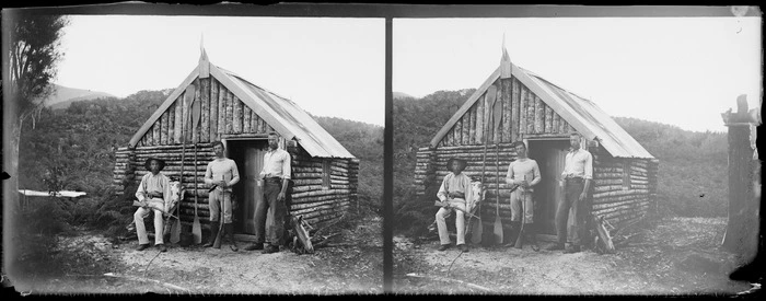 Men, including photographer William Williams (left), outside log cabin set in bush, Southland Region [Fiordland?], including canoe paddles and guns