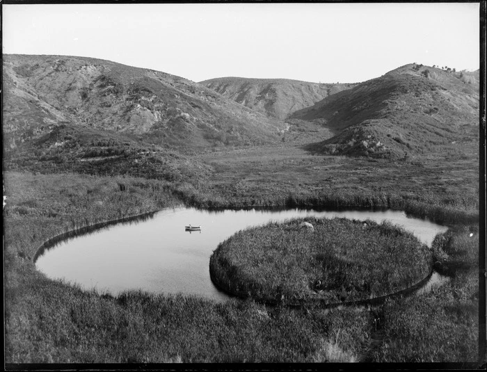 A lake with a circular floating island, including two men in a dinghy, Whakaki, Wairoa District